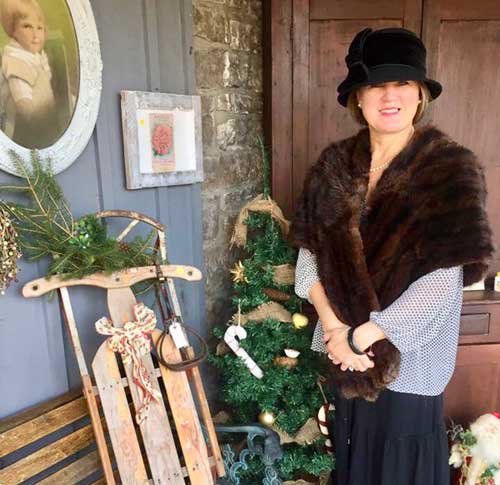 smiling woman wearing hat and fur scarf in front of building with portrait and sled