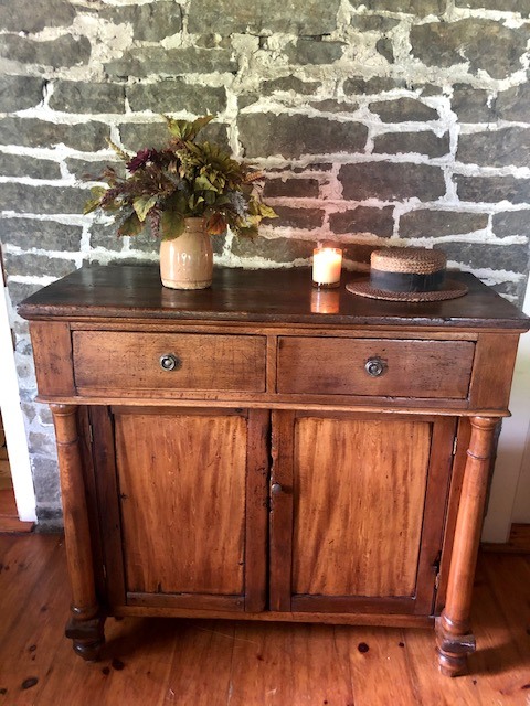 brown wooden cabinet with 2 top drawers, plant in a vase, candle and a hat on top of it.
