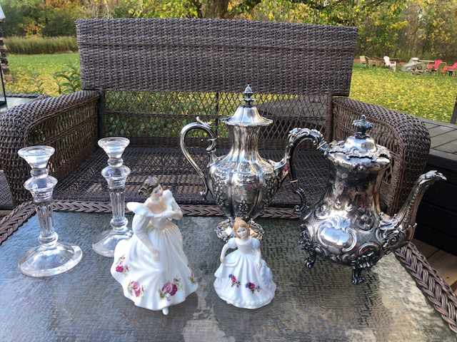 two porcelain figurines, two crystal candlestick holders and two silver teapots on a glass and wicker table in front of a wicker couch