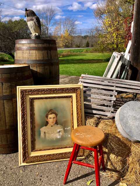 milking stool in front of a portrait of a woman which is leaning against two large wooden barrels with an owl statue on the right barrel
