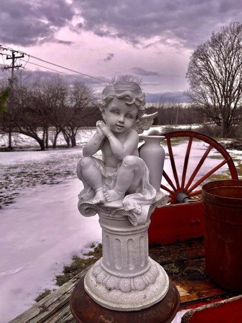 White cherub statue on a pedestal with a snowy field and red wagon behind.