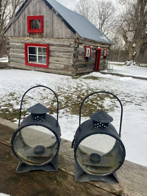 Two black lanterns on a wooden railing with a rustic cabin in a snowy yard behind.