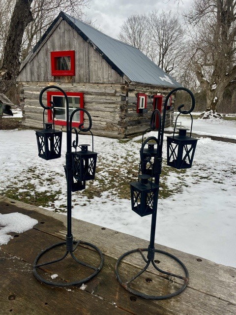 Black metal lantern holders with hanging lanterns in front of a rustic cabin in a snowy yard.