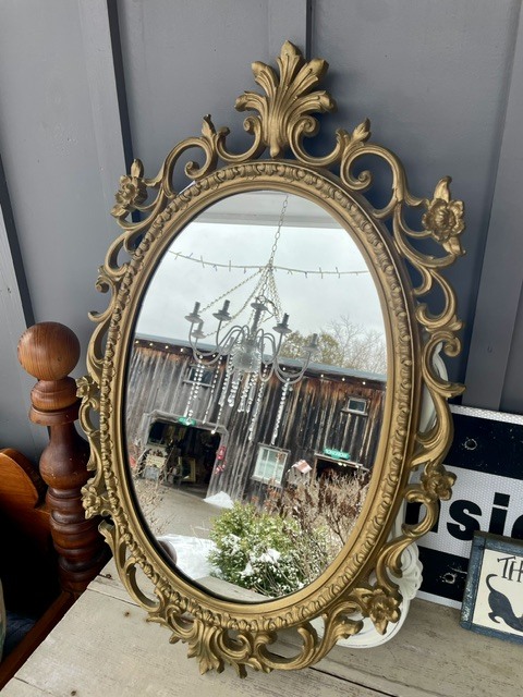 Ornate gold-framed mirror reflecting a barn and chandelier outdoors.