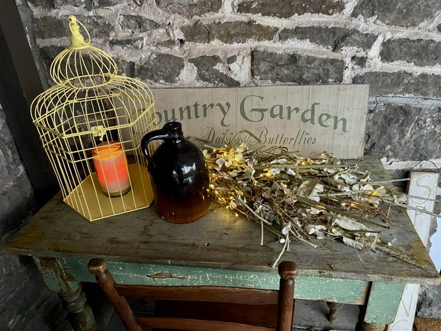 Vintage table with a candle, jug, dried branches, and a decorative garden sign.